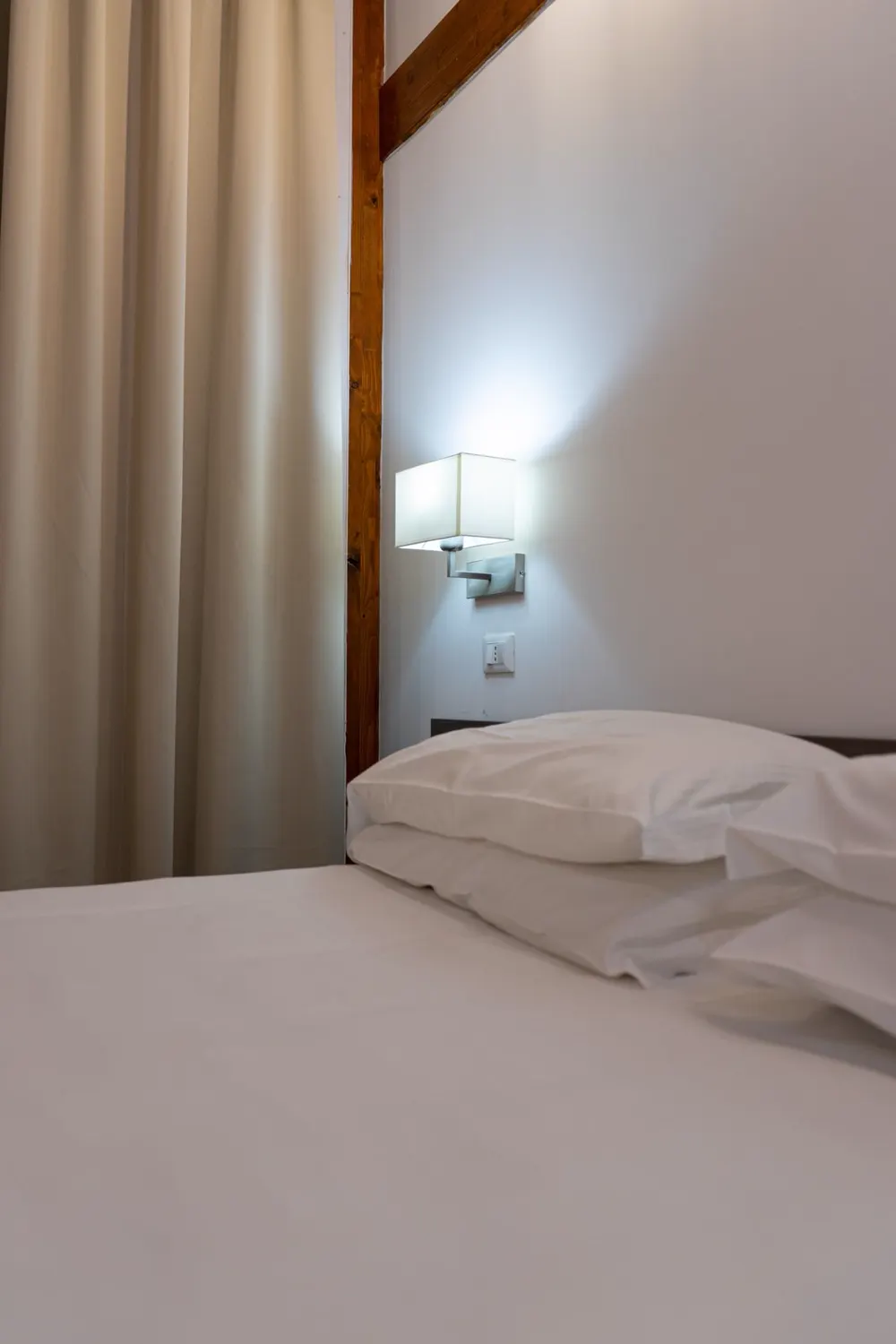 Frisch bezogenes Bett mit Wandleuchte vor hellem Vorhang